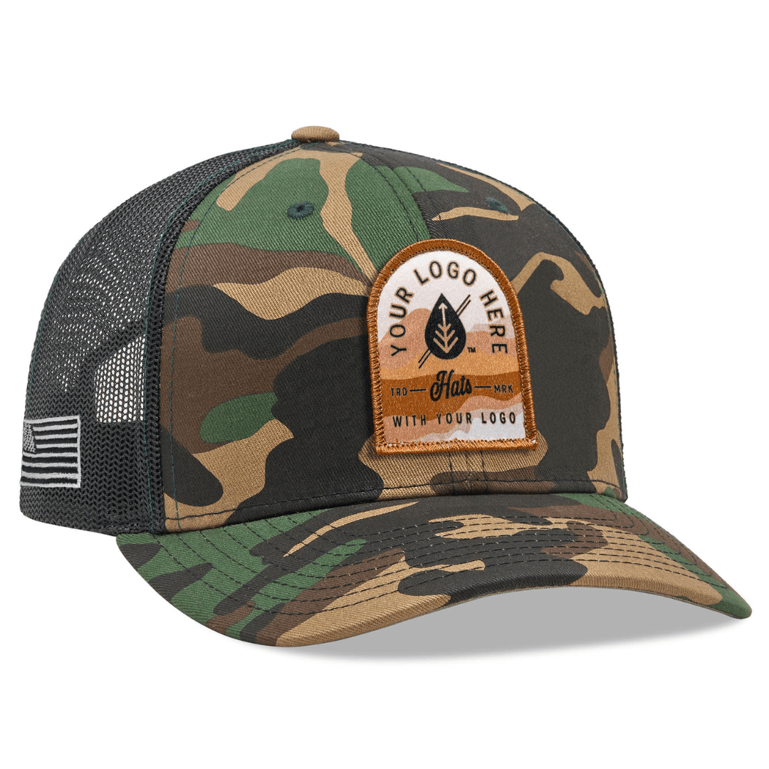 The Richardson 112P Printed Trucker Hat by Richardson is a camouflage cap with a black mesh back, featuring white embroidered text Custom Your Logo Here and an American flag patch on the brims side, perfect for branded or company logo hats.