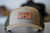 The tan and olive green Richardson 112FP trucker hat highlights a brown leather patch with DEKNI MFG CO. and a leaf logo. With its reflective quality, this custom logo hat merges style and identity seamlessly.